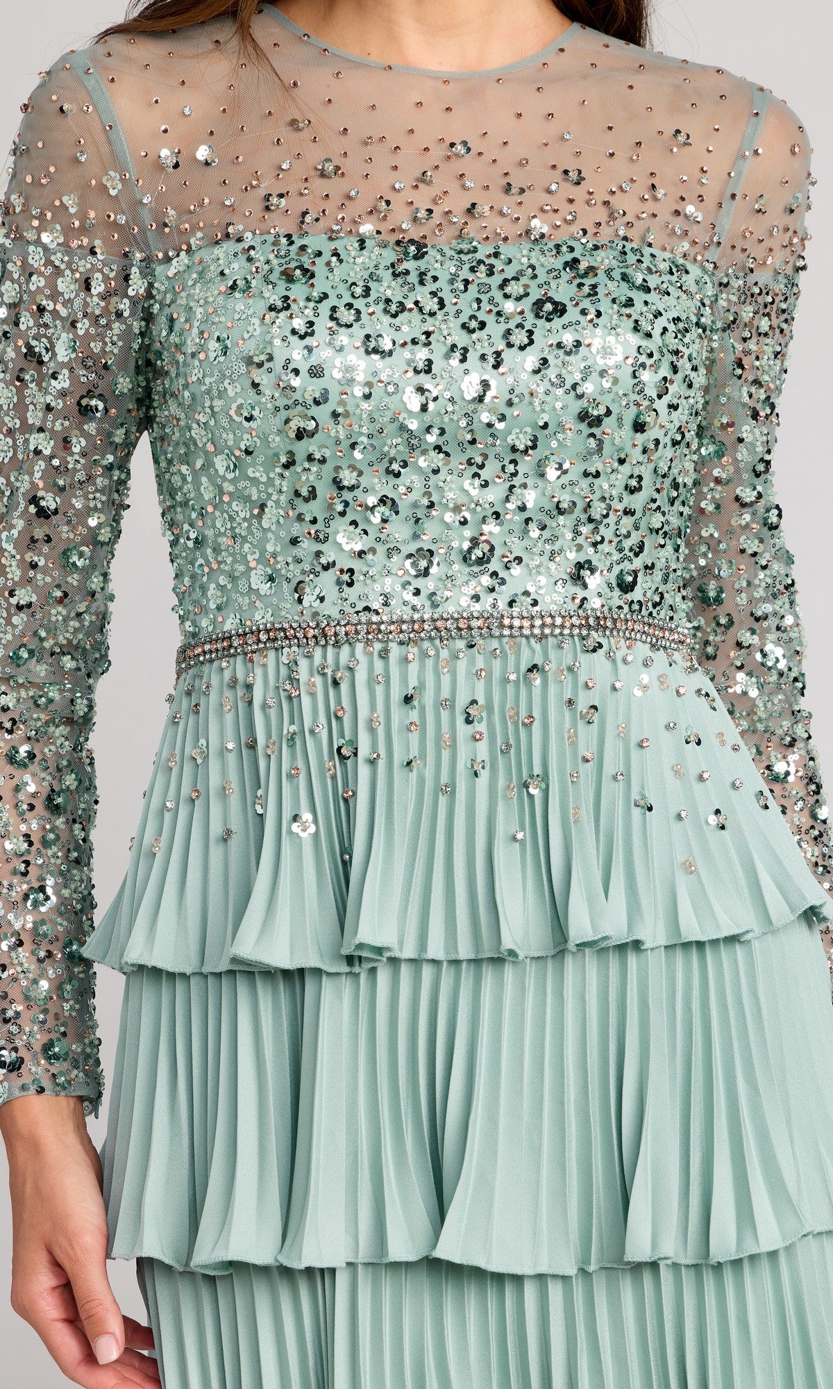 A woman wearing the Mac Duggal 11903 long formal dress in sage green, which features a sequined bodice, sheer long sleeves, and a layered pleated skirt.